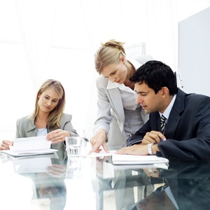 A business woman and business man sit at a glass table looking at paperwork. A third professional, leans over the table and points her finger at a particular part of a document. 